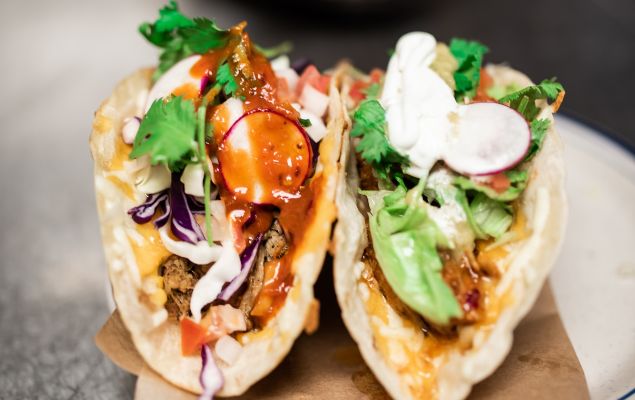 A close-up photo of two tacos bursting with veggies, meat and sauce