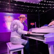 Man dressed in a white sparkly outfit playing a white grand piano.