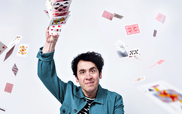 A photo of a man holding a deck of cards above his head and the cards flying all around him.