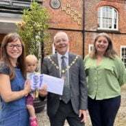 Female holding a baby is presented with an envelope by Mayor and a female wearing a green blouse.