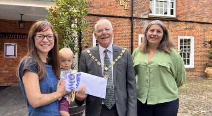 Female holding a baby is presented with an envelope by Mayor and a female wearing a green blouse.