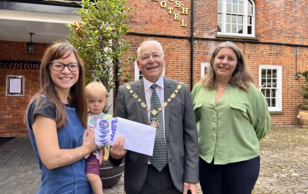 Female holding a baby is presented with an envelope by Mayor and a female wearing a green blouse.