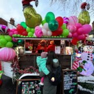 Female with holding a child looking at a Grinch-themed mobile bar covered in pink, green and white balloons.