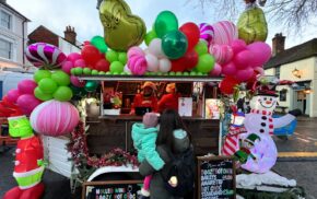 Female with holding a child looking at a Grinch-themed mobile bar covered in pink, green and white balloons.