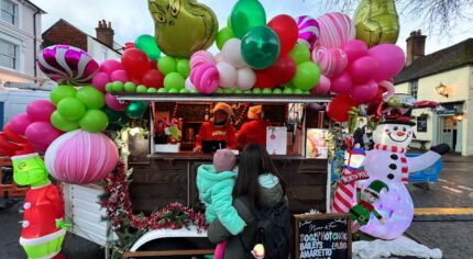 Female with holding a child looking at a Grinch-themed mobile bar covered in pink, green and white balloons.