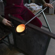 A person is sitting in a work bench, they are using a wooden tool to shape a molten hot piece of glass on the end of a metal rod. The glass is so hot it is glowing orange.