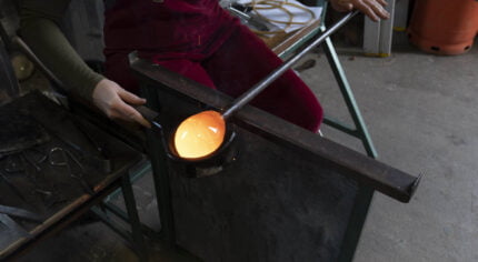 A person is sitting in a work bench, they are using a wooden tool to shape a molten hot piece of glass on the end of a metal rod. The glass is so hot it is glowing orange.