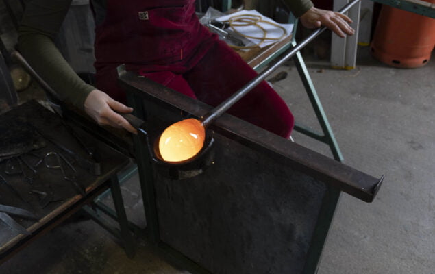 A person is sitting in a work bench, they are using a wooden tool to shape a molten hot piece of glass on the end of a metal rod. The glass is so hot it is glowing orange.