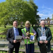 Three people standing in front of wooden fence. The two men on the left and right are holding copies of a brochure. The female in the centre is wearing a high viz jacket and is holding a small dog.