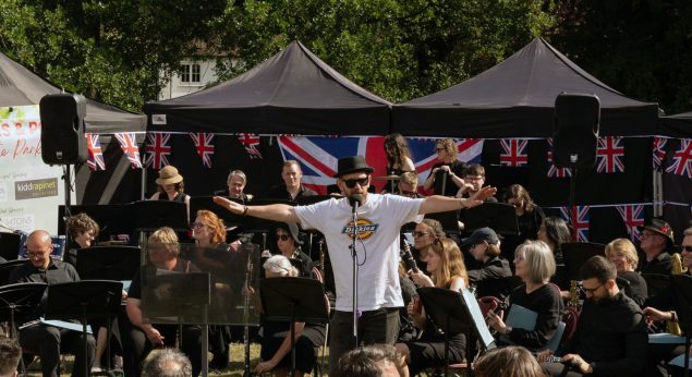 Male singer standing with arms outstretched in front of a crowd, with Union Jack flags in the background.