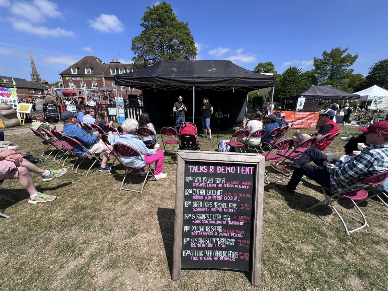 Chalk board advertising details of talks. In the background there are people speaking to a small crowd of people.