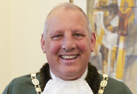 Smiling man dressed in a green Mayoral robe, a white lace jabot and gold chain of office.