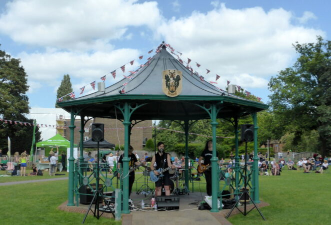 Three males playing electric guitar in a bandstand in a park.