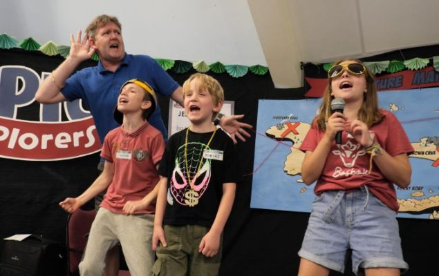 Three children singing. An adult in the background
