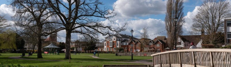 Winter scene of town centre park. Wooden bridge in foreground, trees, buildings and bandstand in background. Blue sky.