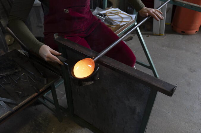 A person is sitting in a work bench, they are using a wooden tool to shape a molten hot piece of glass on the end of a metal rod. The glass is so hot it is glowing orange.