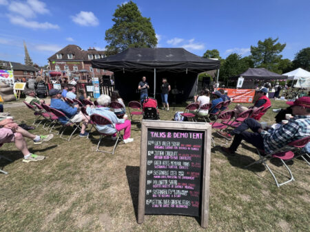 Chalk board advertising details of talks. In the background there are people speaking to a small crowd of people.