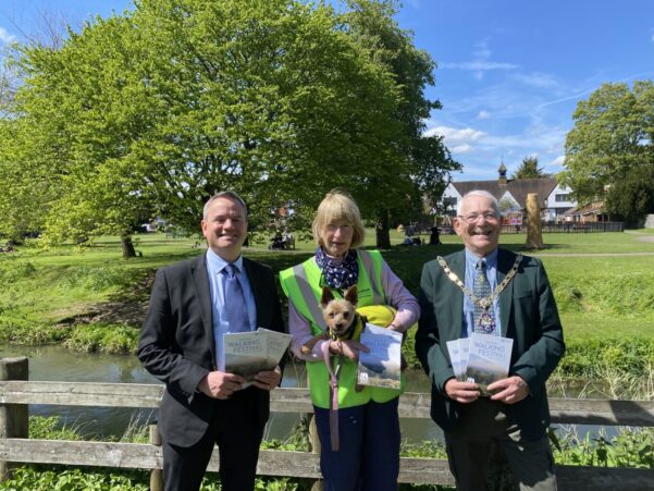 Three people standing in front of wooden fence. The two men on the left and right are holding copies of a brochure. The female in the centre is wearing a high viz jacket and is holding a small dog.