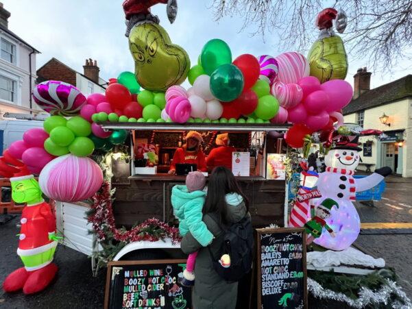 Female with holding a child looking at a Grinch-themed mobile bar covered in pink, green and white balloons.