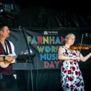 Man playing a banjo and a woman playing a violin on a stage with a World Music Day banner in the background.
