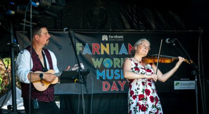 Man playing a banjo and a woman playing a violin on a stage with a World Music Day banner in the background.