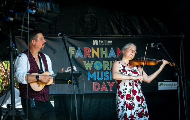 Man playing a banjo and a woman playing a violin on a stage with a World Music Day banner in the background.