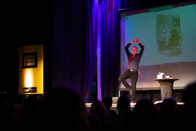 Man on a stage standing on one leg with his arms above his head.