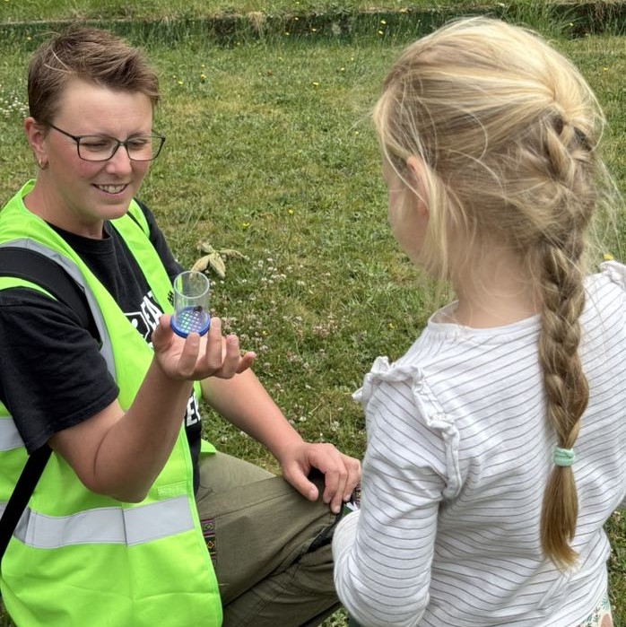 Person in a high viz waistcoat showing a young girl a bee in a glass jar.