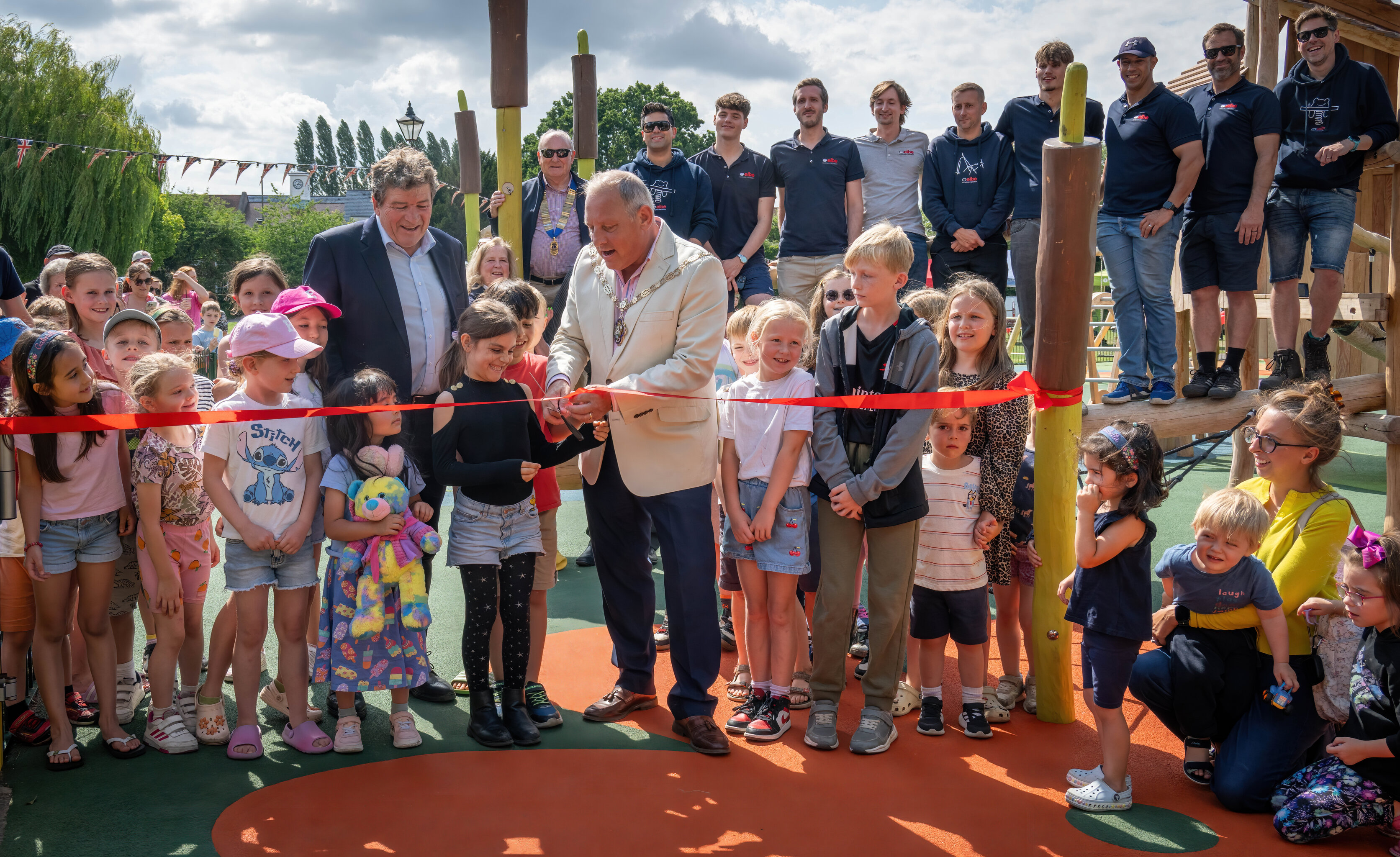 A crowd of adults and children watch as the Mayor and children cut a ribbon to open a new playground.