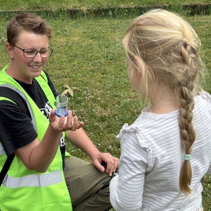 Young girl looking at a bee in a specimen jar held by a woman in a high viz jacket.