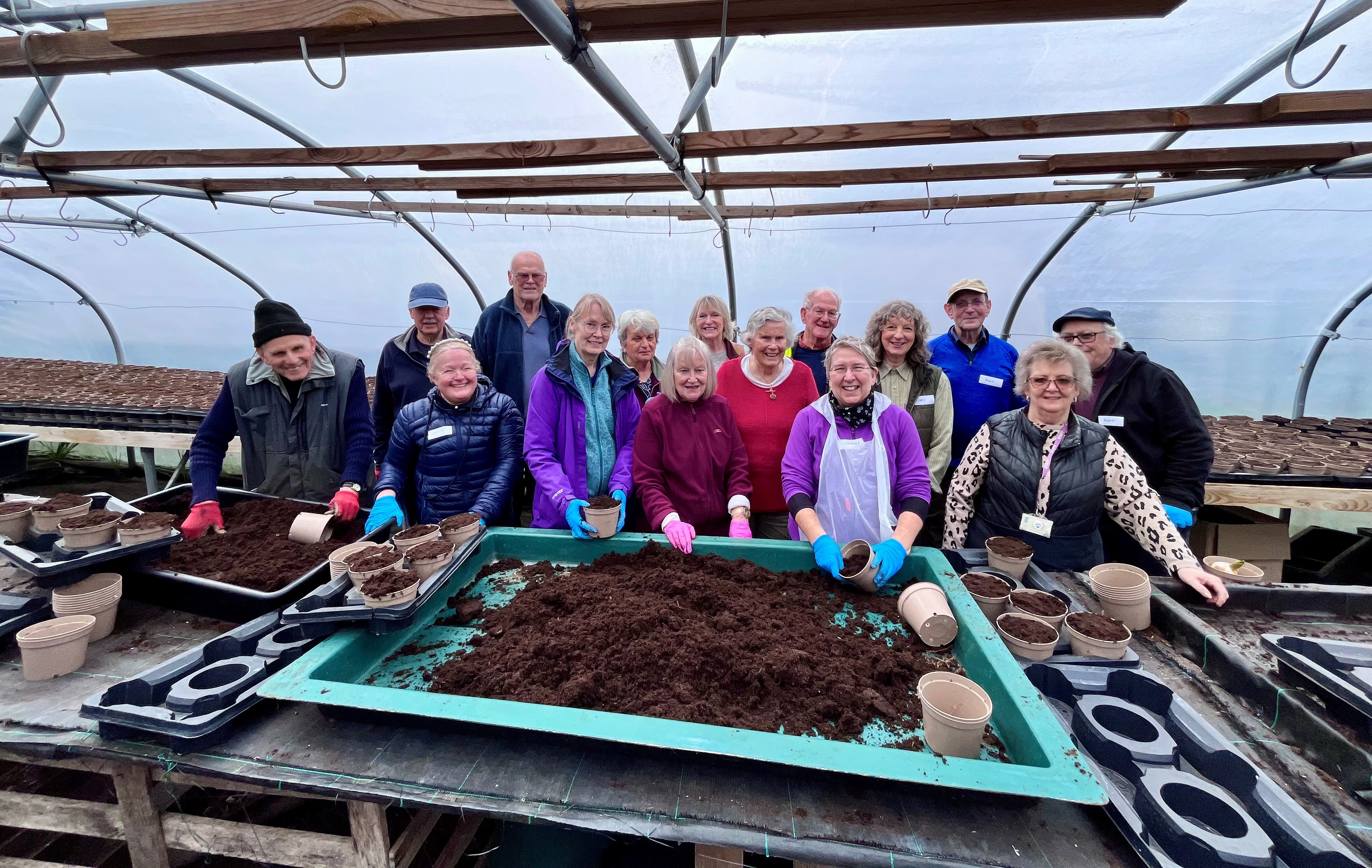 Group of people in a greenhouse, standing behind a big tray of compost.