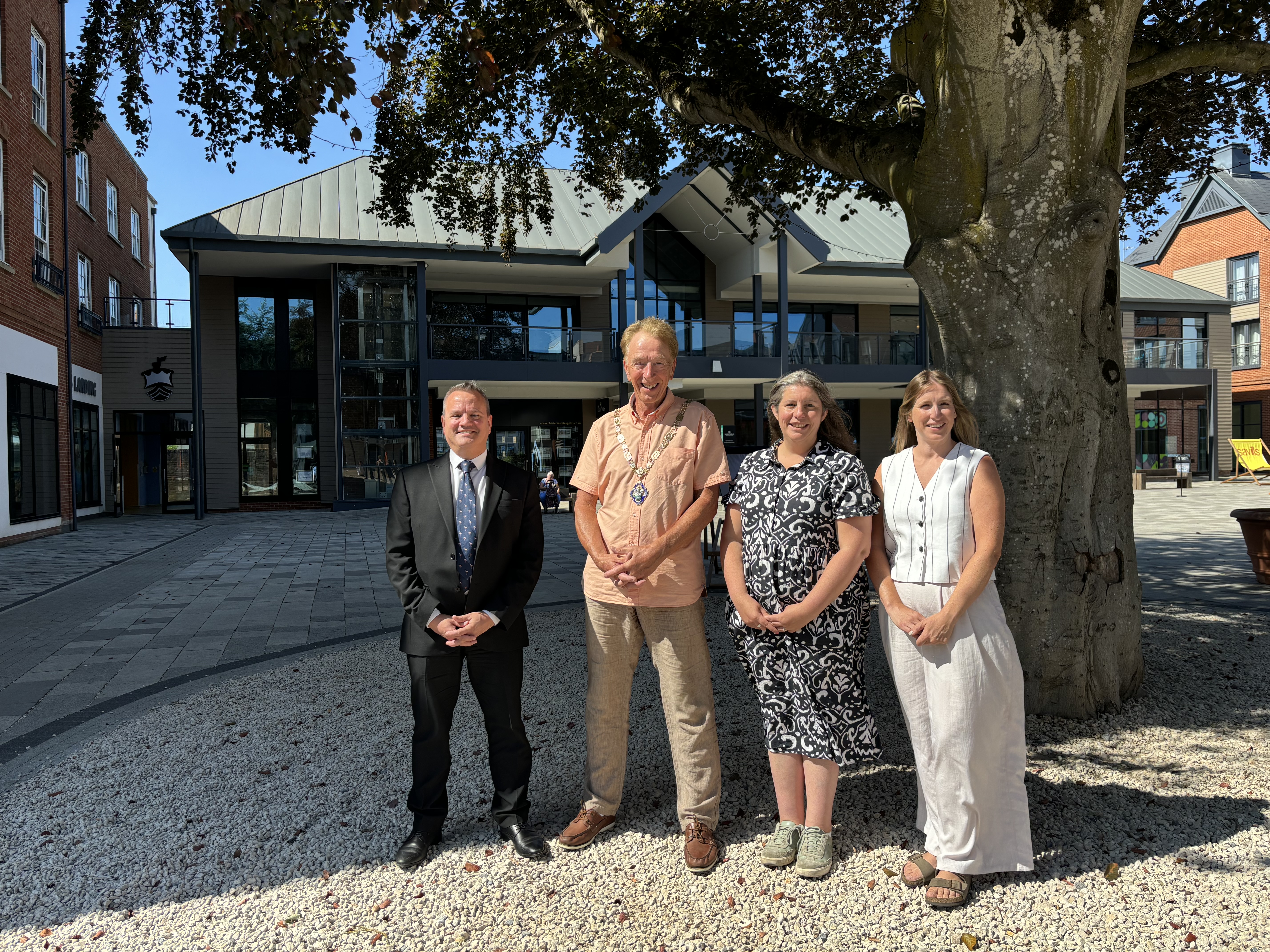 Four people standing under a tree with buildings in the background.