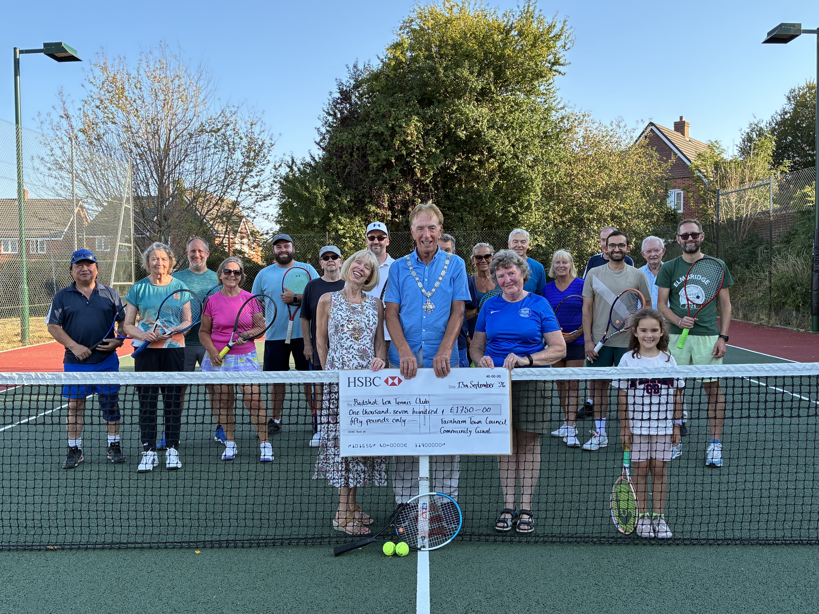 Group of people standing on a court behind a tennis net. The person at the front is holding a large cheque.
